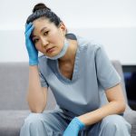 Tired nurse in blue scrubs resting with hand on head, expressing exhaustion.