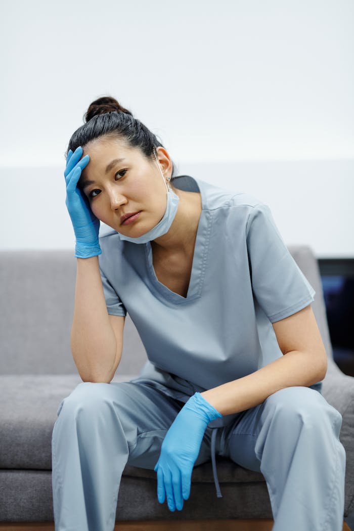 Tired nurse in blue scrubs resting with hand on head, expressing exhaustion.